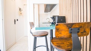 A kitchen area with a countertop, a coffee machine, and a toaster. There are two chairs, one with a wooden seat and backrest, and the other with a metal frame and wooden seat. A white door is visible to the left. - Studio for 2 People - Hotel de Ville - Marais - Paris 04 - Central (Paris)