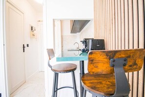 Eine Küchenzeile mit Arbeitsplatte, einer Kaffeemaschine und einem Toaster. Zwei Stühle sind vorhanden, einer mit Holz-Sitzfläche und -Lehne, der andere mit Metallgestell und Sitzfläche aus Holz. Eine weiße Tür ist links im Bild zu sehen.