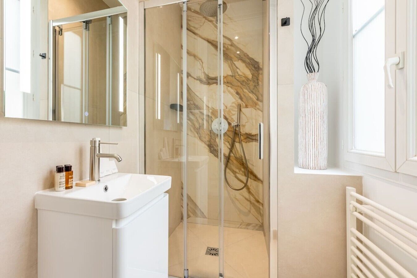 Ein modernes Badezimmer mit einer gläsernen Duschkabine, einem weißen Waschbecken mit Spiegel darüber und einem Heizkörper rechts.