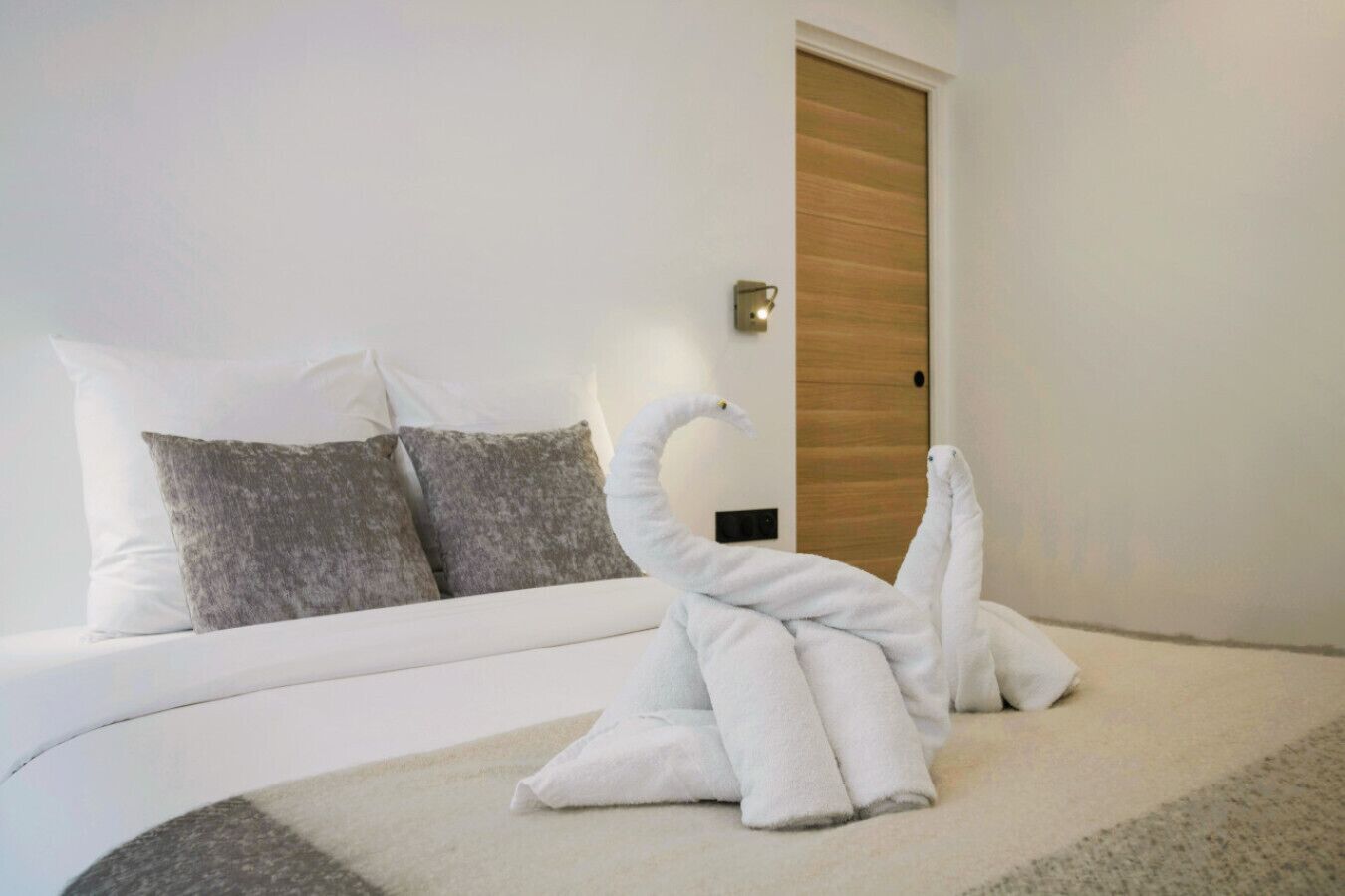 Quarto de hotel com cama, almofadas, porta de madeira e luz fixada na parede.