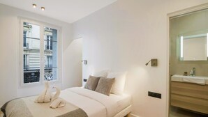 A bedroom with a bed, pillows, a bedside table, a mirror, and a window with blinds. - Premium Suites - Arc de Triomphe - Trocadéro (Paris)