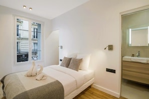 A bedroom with a bed, pillows, a bedside table, a mirror, and a window with blinds. - Premium Suites - Arc de Triomphe - Trocadéro (Paris)