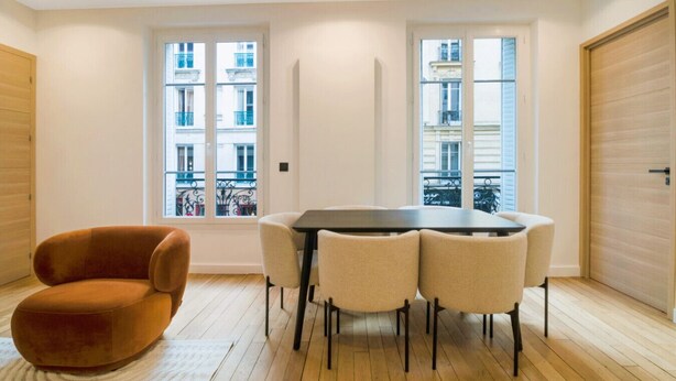 A modern interior with a dining table surrounded by chairs, a single sofa, and large windows with a view of buildings. - Premium Suites - Arc de Triomphe - Trocadéro (Paris)