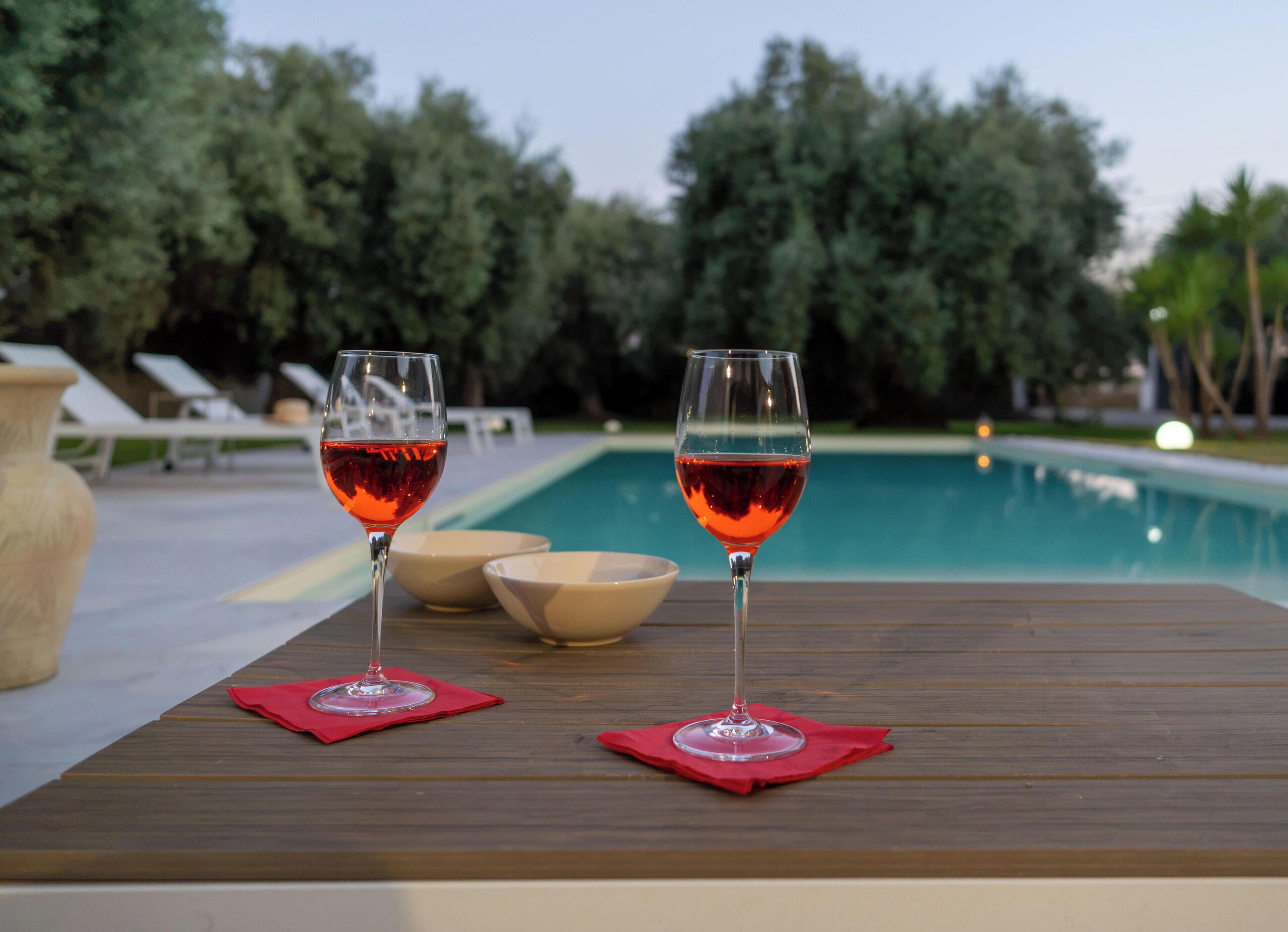 Deux verres de vin rouge sur une table en bois, près d’une piscine, avec des chaises longues et une vue sur les arbres.