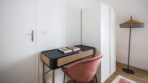 Een kleine kamer met een bureau, stoel en een vloerlamp.
