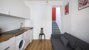 Een ruimtelijk ingerichte leefruimte met kitchenette, een wasmachine, een grijze bank en een trap naar een verdieping.