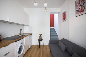 Ein kompakter Wohnbereich mit Küchenzeile, einer Waschmaschine, einem grauen Sofa und einer Treppe zu einer oberen Ebene.