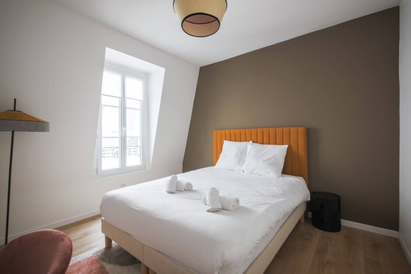A hotel room with a bed, a window, a floor lamp, and a trash bin.