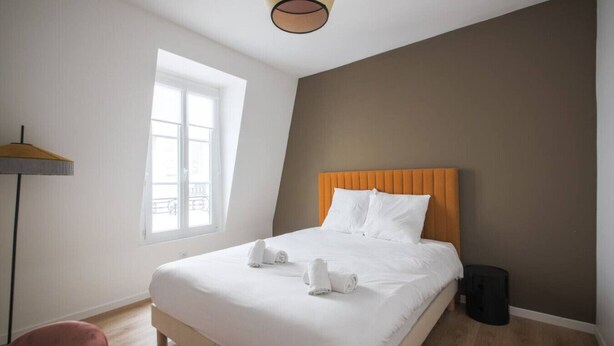 Een hotelkamer met een bed, een raam, een vloerlamp en een prullenbak.
