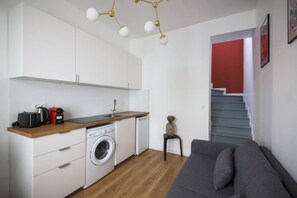 Eine kompakte Küche mit Waschmaschine, Kaffeemaschine und Toaster. Ein graues Sofa und ein kleiner Tisch mit einer Vase sind zu sehen. Eine Treppe mit rotem Akzent ist sichtbar.