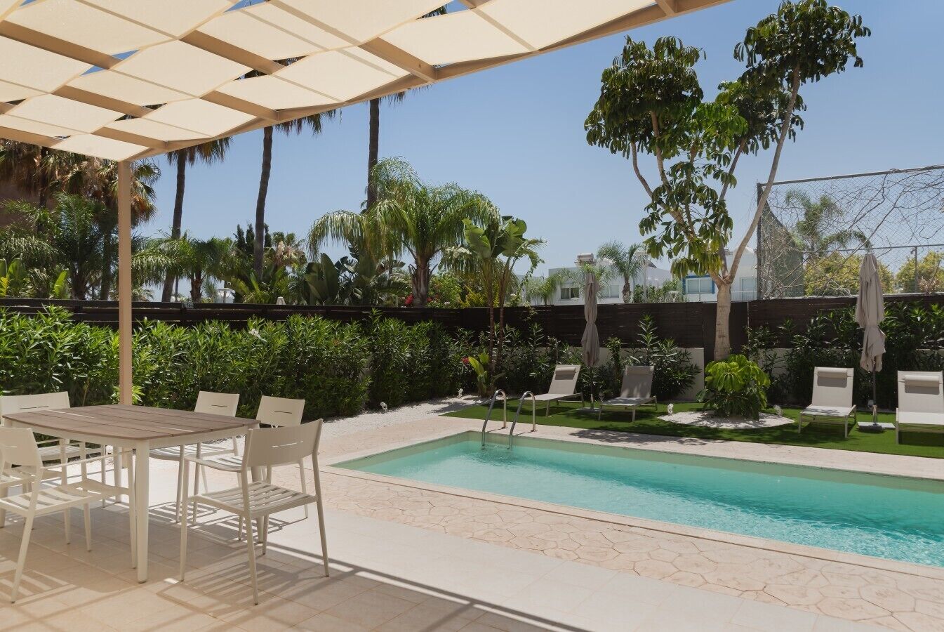 A pool area with a table and chairs, surrounded by greenery and a fence.