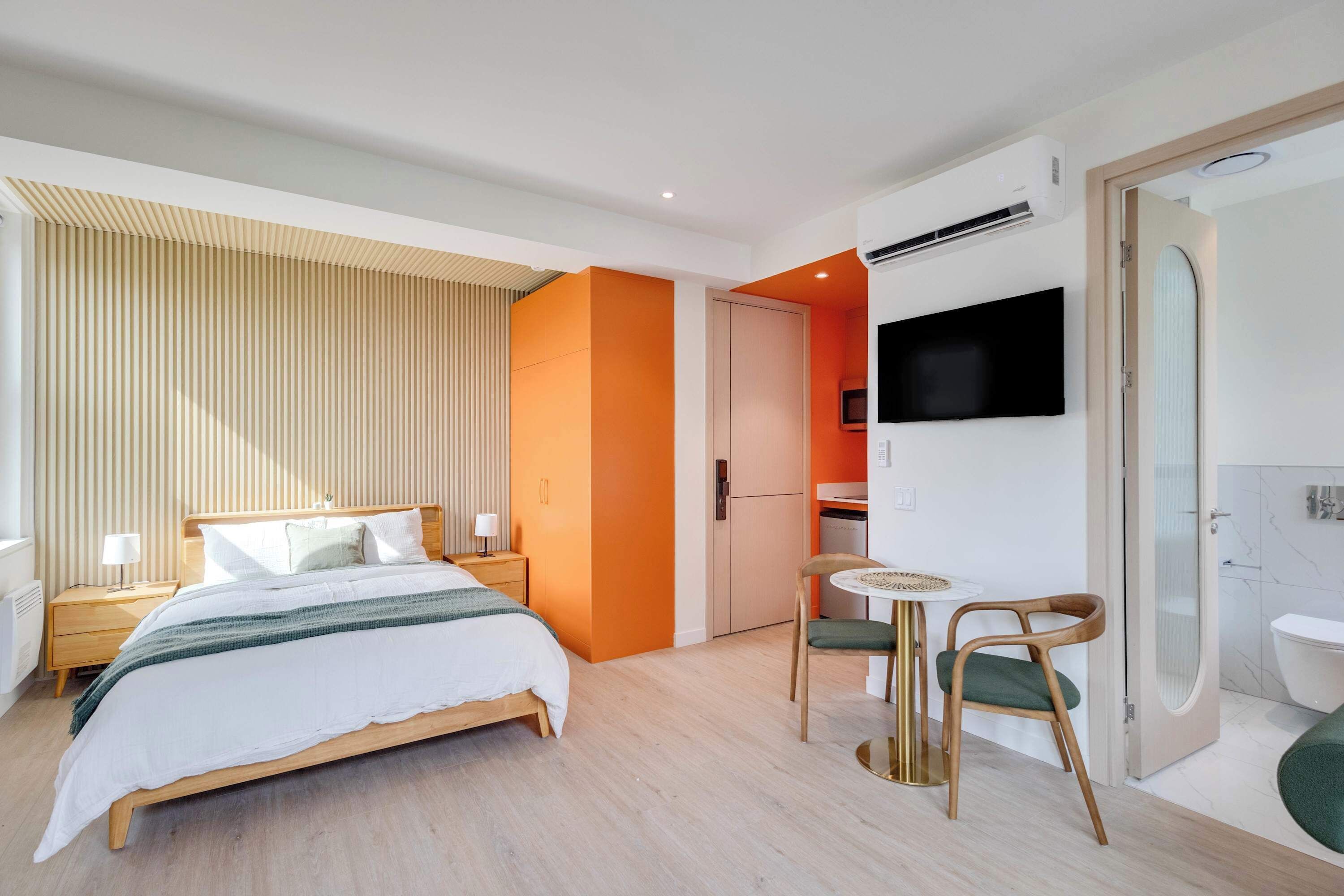 A modern hotel room with a bed, bedside tables, a TV, a small table with chairs, and a bathroom with a toilet and shower.