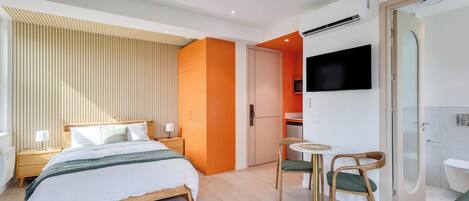 A modern hotel room with a bed, bedside tables, a TV, a small table with chairs, and a bathroom with a toilet and shower.