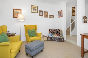 Een zithoek met een gele fauteuil, een grijze poef en een vloerlamp. Er hangen fotolijsten aan de muur en een trap is zichtbaar op de achtergrond.