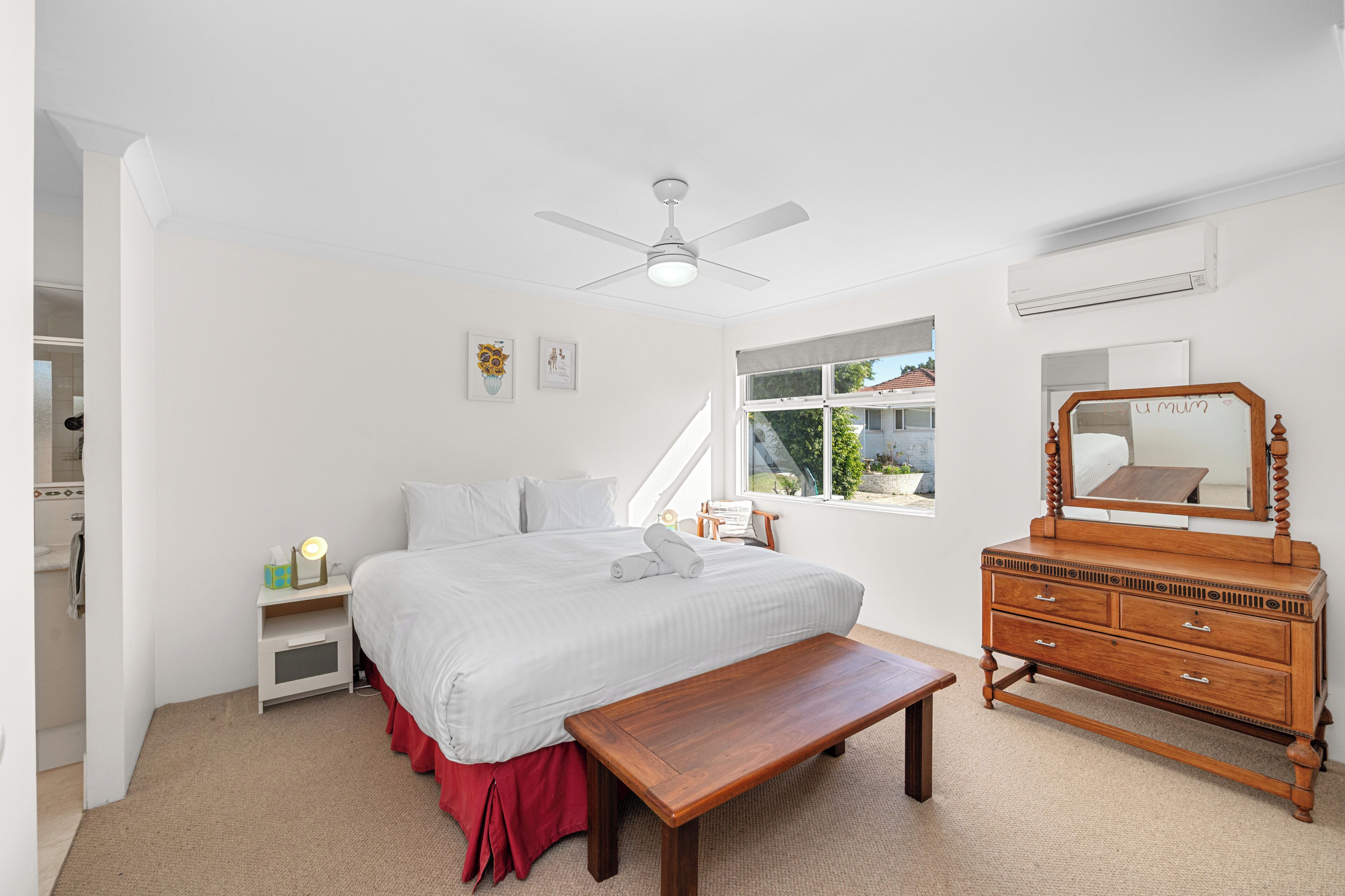 A bedroom with a bed, a wooden dresser, a bench, a ceiling fan, and a window with a view of the outside.