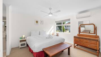 A bedroom with a bed, a wooden dresser, a bench, a ceiling fan, and a window with a view of the outside.