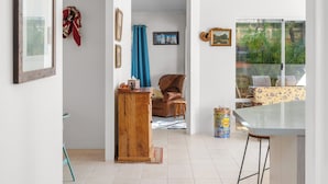 Un'ampia stanza con pavimento piastrellato, un armadio in legno, una poltrona in pelle marrone, un tavolo da pranzo con sedie e una finestra vista su del verde.