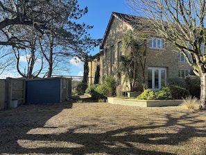 A stone building with a gravel driveway, a wooden fence, and a variety of shrubs and trees.