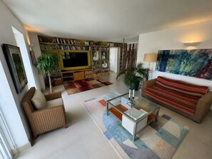 A living room with a sofa, armchair, coffee table, and a television. There is a bookshelf filled with books and a painting on the wall. - Outstanding contemporary country house, close to Bath with stunning views. (Bath)