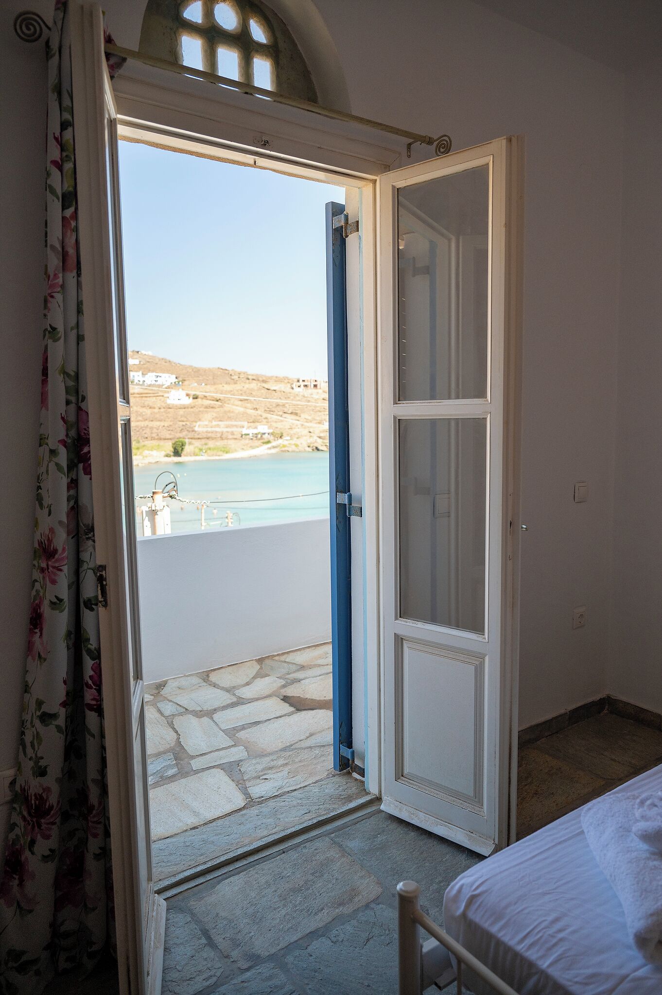Una habitación con cama, una puerta que da a un balcón con vista al mar y una ventana con cortina.