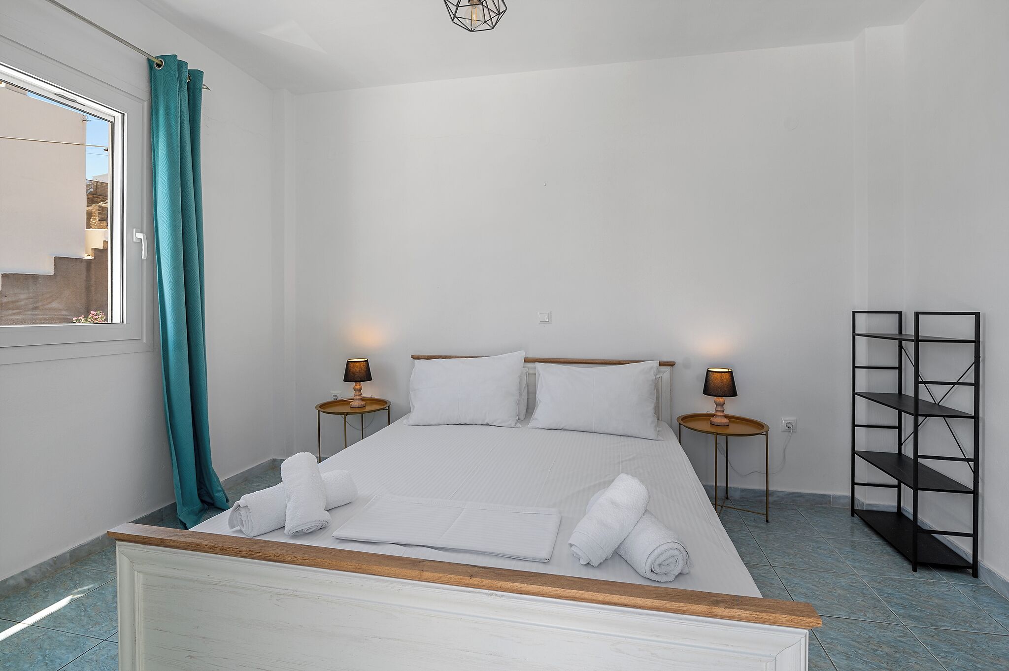 A bedroom with a bed, two bedside tables, a wall-mounted shelf, and a window with curtains.