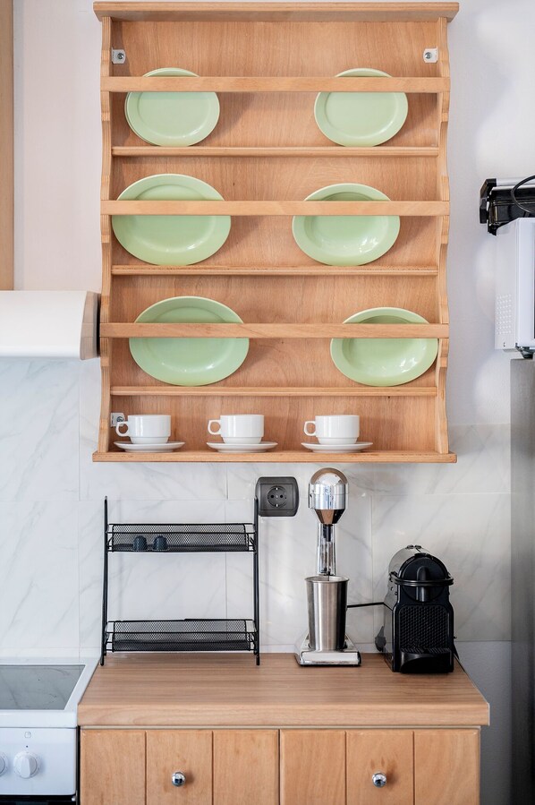 Una cocina con un estante de madera que contiene platos verdes y tazas blancas, una cafetera y un hervidor sobre la encimera.