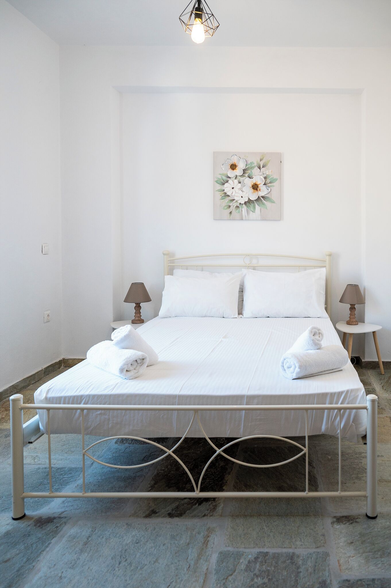 A neatly made bed with white linens and towels, a wall-mounted light fixture, and a floral painting above the bed.
