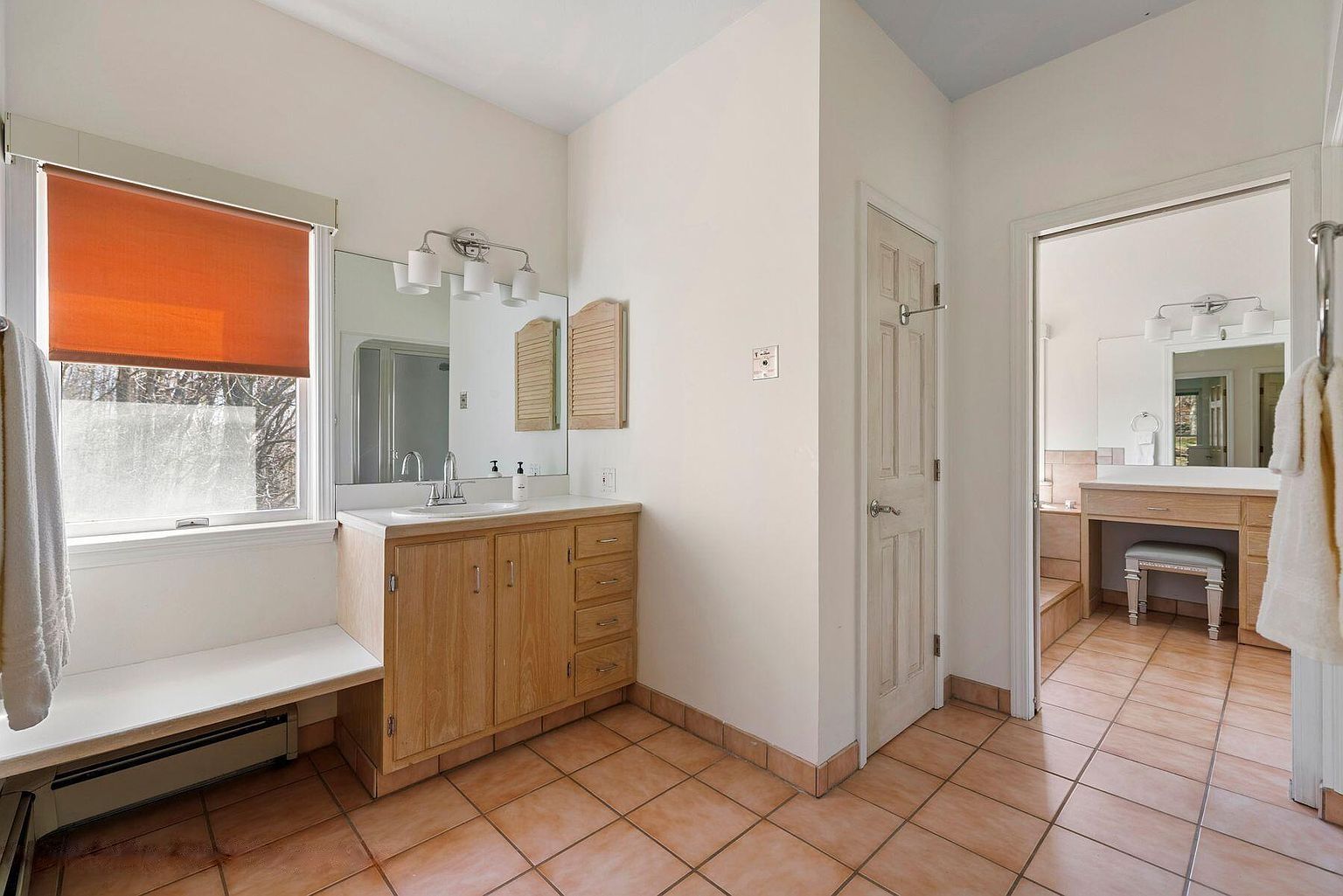 Une salle de bain avec un meuble-lavabo en bois, un grand miroir et une fenêtre avec un store orange.