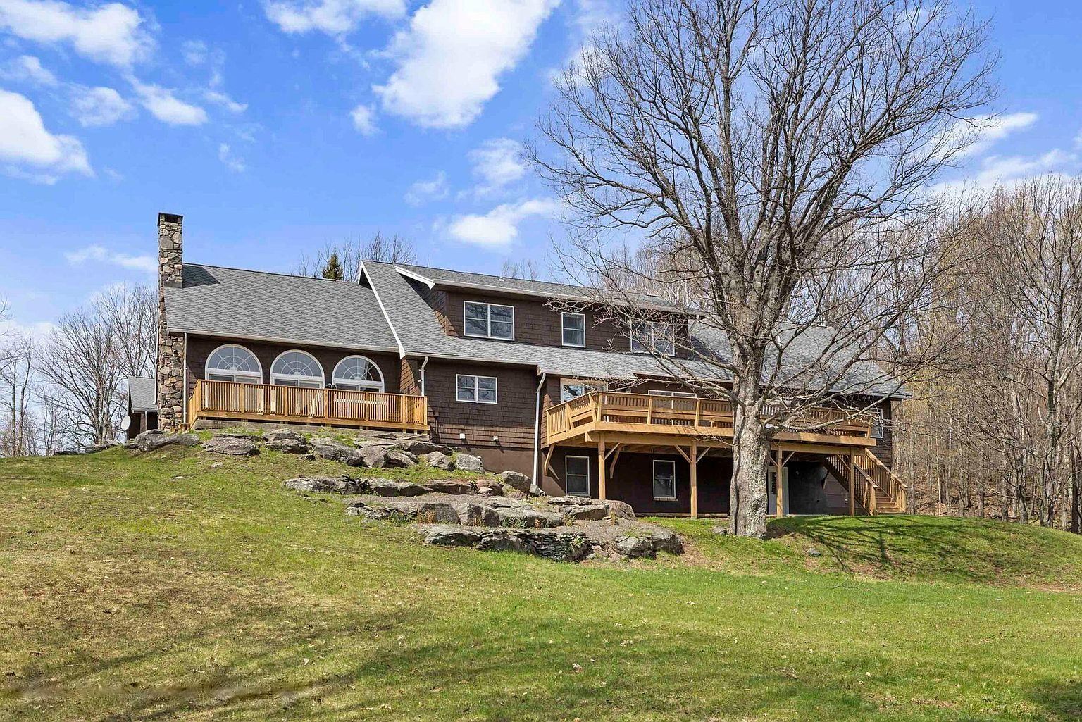 A two-story house with a stone chimney, wooden deck, and a large tree in the background.
