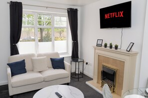 A living room with a white sofa, black curtains, a fireplace, a television mounted on the wall, and a small table with a remote control. - Boutique 2-Bed Apartment in Wolverhampton Centre (Wolverhampton)