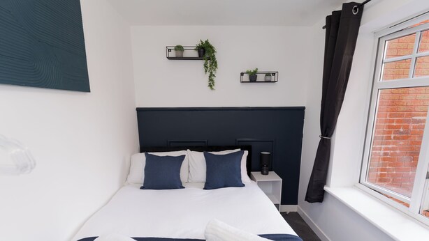 A neatly made bed with pillows and a blanket, a bedside table with a lamp, and a window with curtains. - Boutique 2-Bed Apartment in Wolverhampton Centre (Wolverhampton)