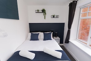Ein ordentlich bezogenes Bett mit Kissen und Decke, ein Nachttisch mit Lampe und ein Fenster mit Vorhängen.