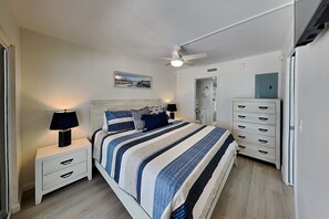 A bedroom with a bed, nightstands, a dresser, a ceiling fan, and a door leading to another room. - Pointe South 401 | Resort Views + Pool (Fort Myers Beach)