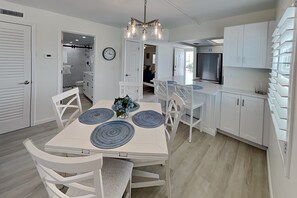 A modern kitchen with white cabinetry, a dining area with a white table and chairs, and a built-in TV above the counter. - Pointe South 401 | Resort Views + Pool (Fort Myers Beach)