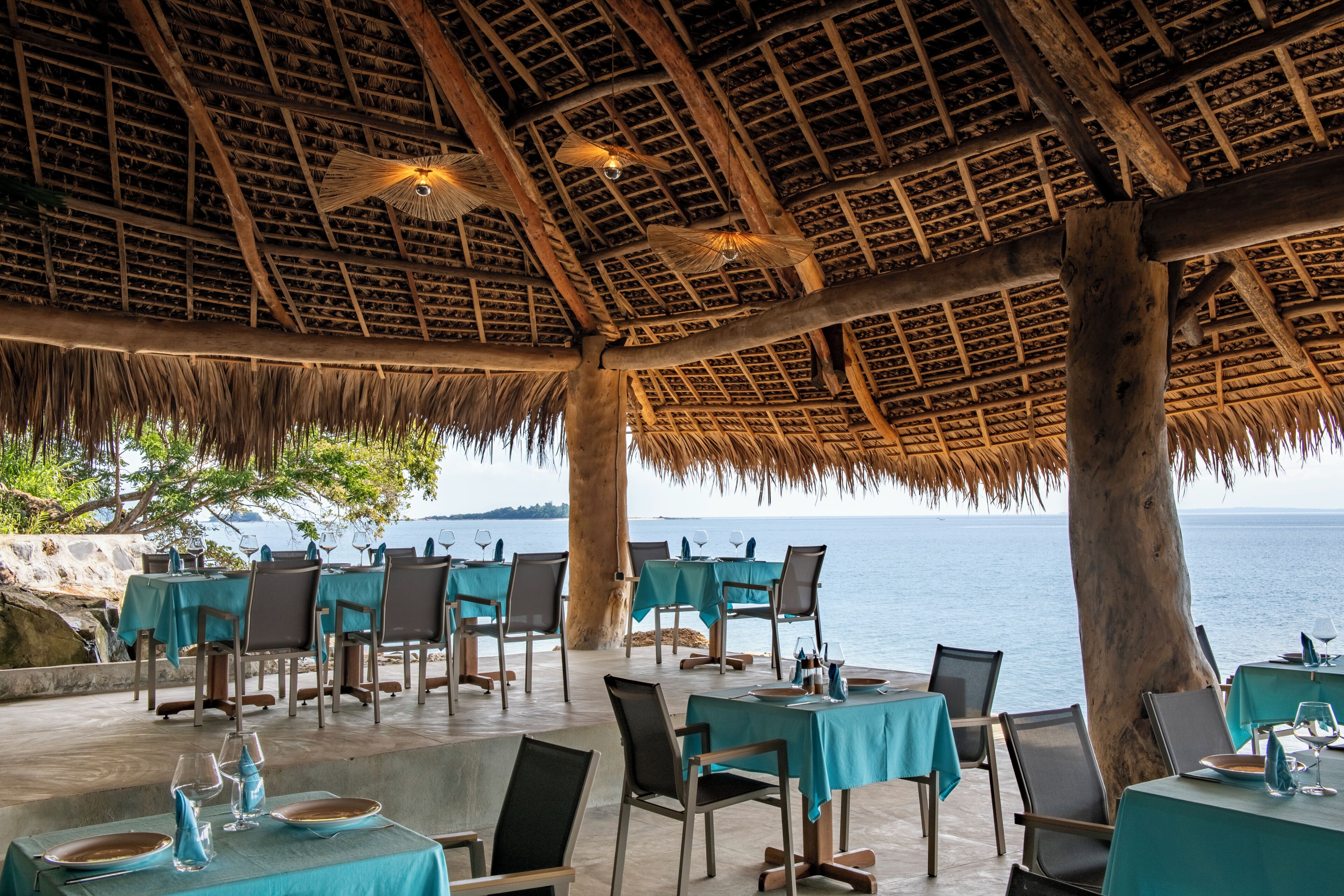 An outdoor dining area with tables and chairs, a thatched roof, and a view of the sea.