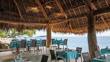 An outdoor dining area with tables and chairs, a thatched roof, and a view of the sea.