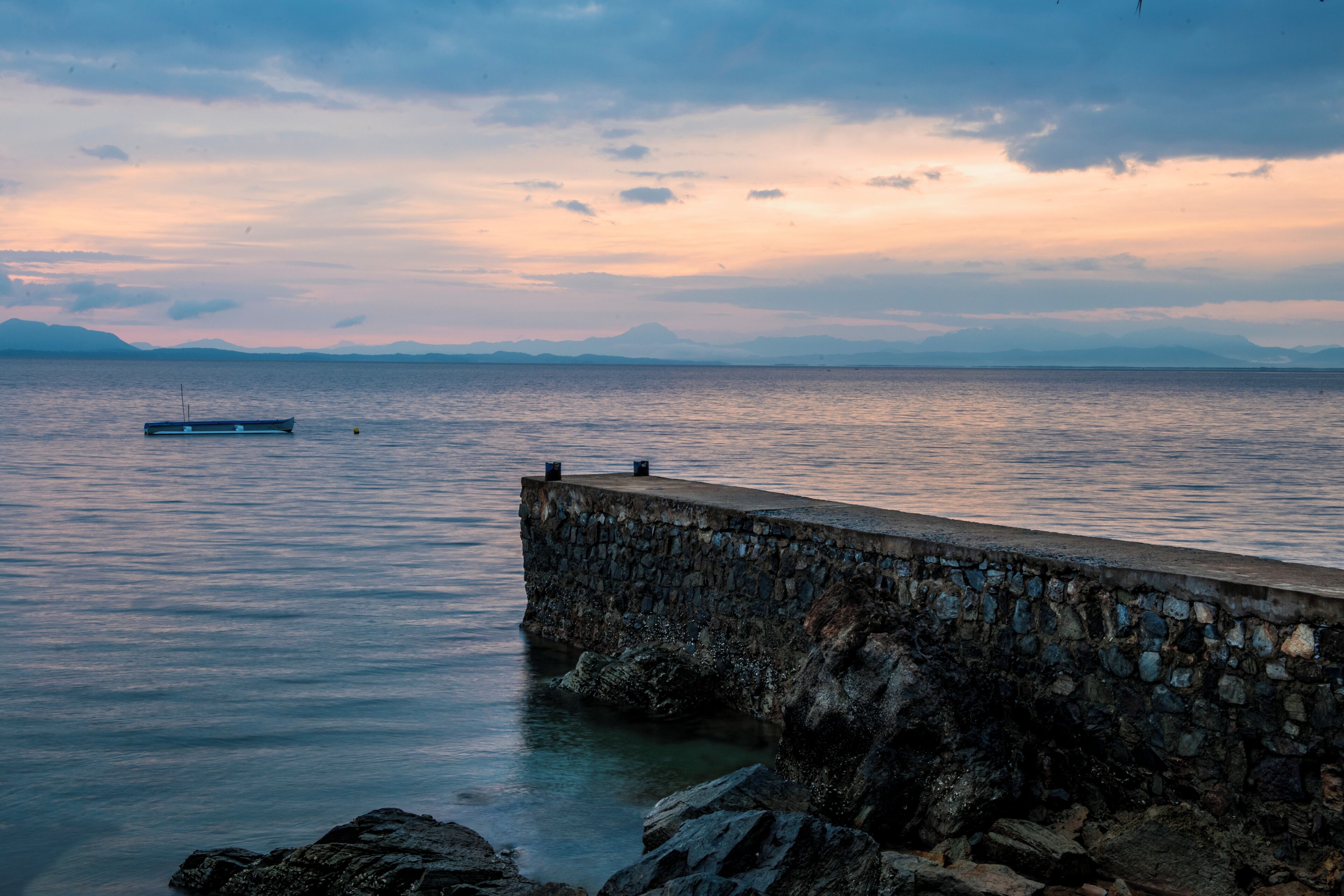 Un muelle de piedra se adentra en aguas tranquilas, con un barco anclado a lo lejos y montañas al fondo.