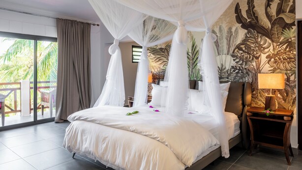 A room with a four-poster bed, a balcony with a view of palm trees, and a mural of tropical leaves on the wall. - Ankomba Hotel (Nosy Komba)