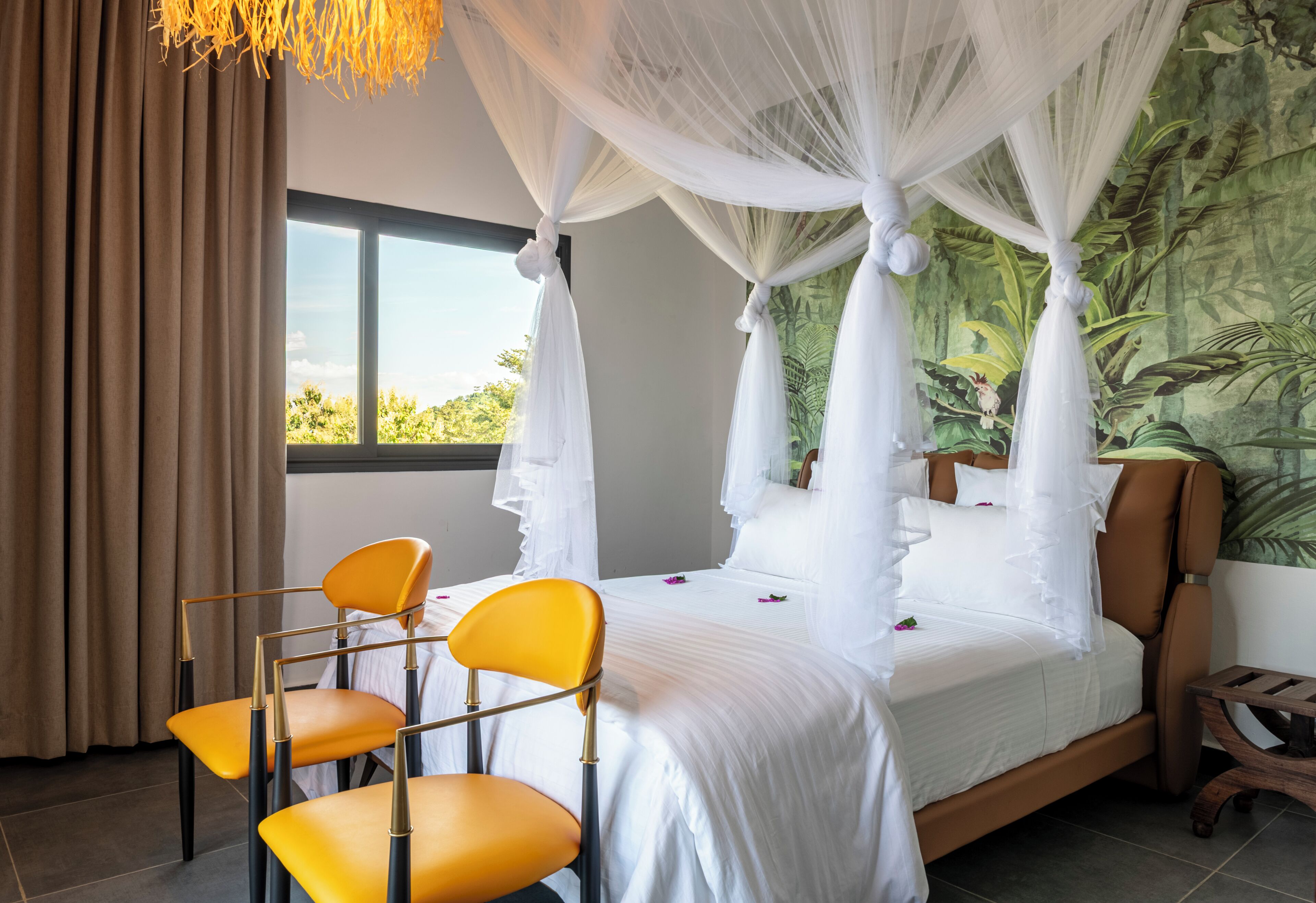 Uma cama com dossel e cortinas, roupa de cama branca, duas cadeiras amarelas e vista para o verde através de uma janela.