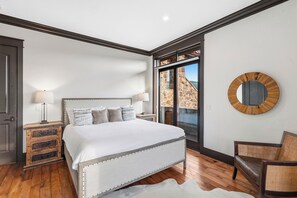 Ein Schlafzimmer mit einem großen Bett, einem Holznachttisch, einem Sessel und Blick durch eine Glastür.