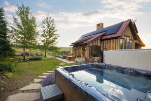 Ein Whirlpool mit Holzterrasse, eine Terrasse mit Gartenmöbeln und ein Haus mit Steinofen.