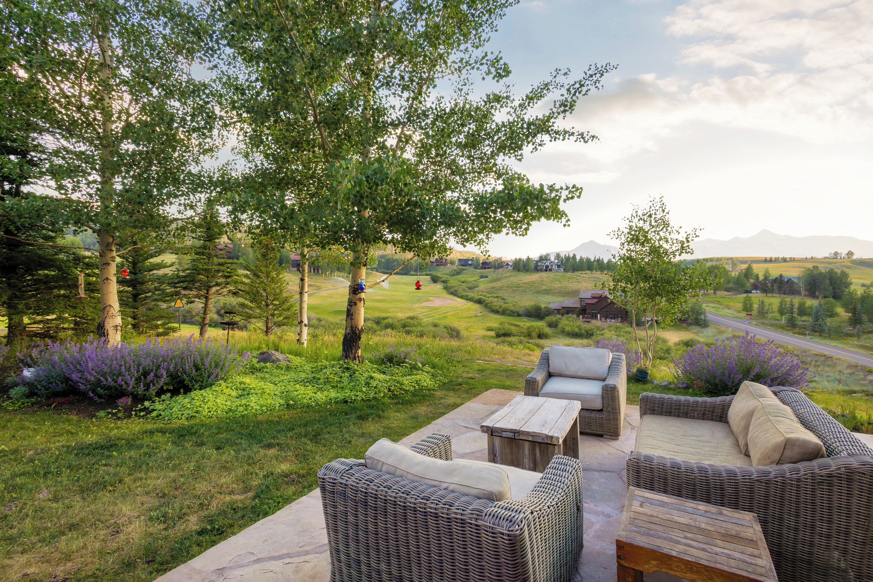 An outdoor seating area with wicker furniture, a stone table, and a view of a rural landscape with trees and a road.
