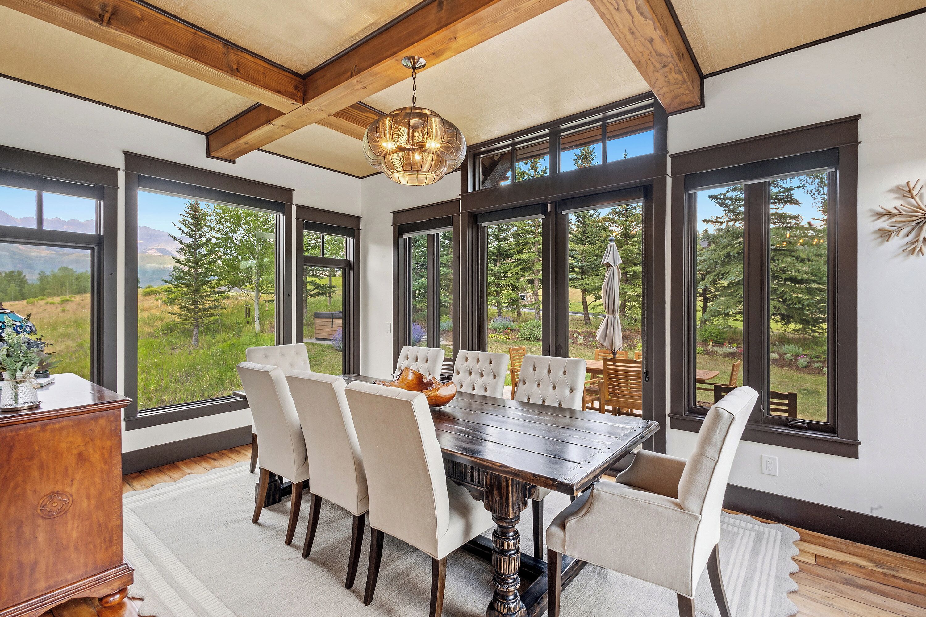 A dining room with a large table and chairs, large windows with a view of greenery and mountains, and a wooden ceiling with exposed beams.