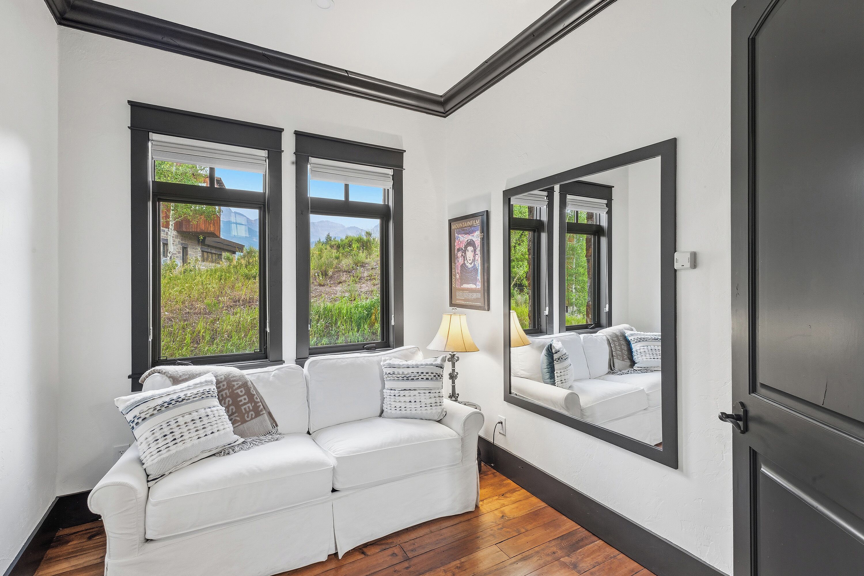 A room with a white sofa, a large mirror, and two windows with a view of greenery and mountains.