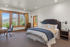 Ein Schlafzimmer mit einem großen Bett, einem Schreibtisch und einem Stuhl. Es gibt Fenster mit Vorhängen, einen Nachttisch und eine Lampe.