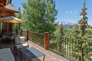 Una terraza de madera con mobiliario exterior, barandilla y vistas a una cadena montañosa.