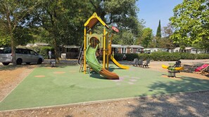 A playground with a slide and swings, surrounded by trees and a parking area. - Lou Cigalou - Mobile Home - Sleeps 6 (La Roque-d'Anthéron)