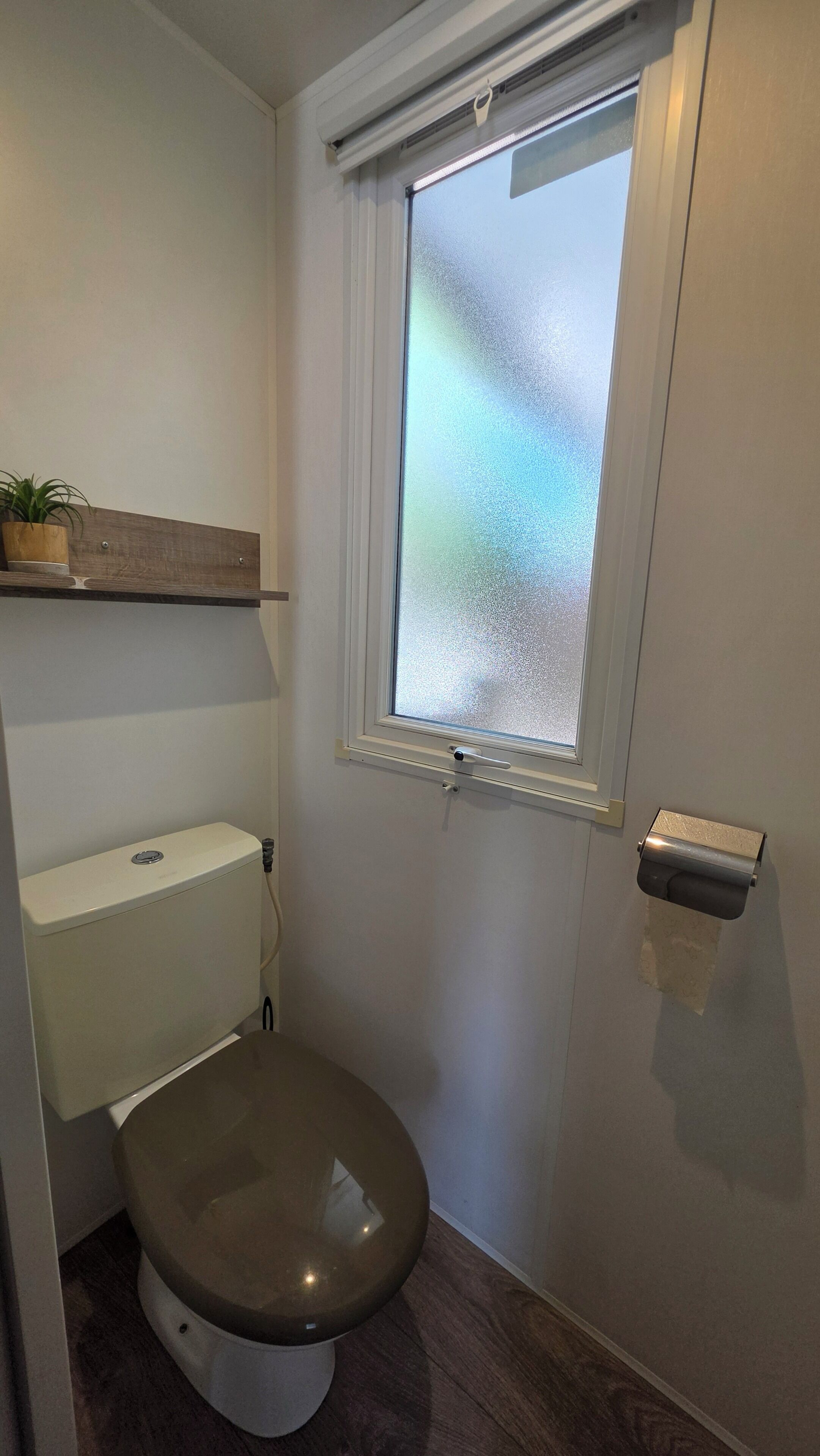 Une salle de bain avec des toilettes, une fenêtre à vitre dépolie et une étagère en bois au-dessus des toilettes.