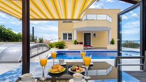 A dining table with breakfast items and drinks, overlooking a swimming pool and a modern building. - Villa 'Rocco-Salzwasserpool-Split' with Wi-Fi (Kaštel Gomilica)