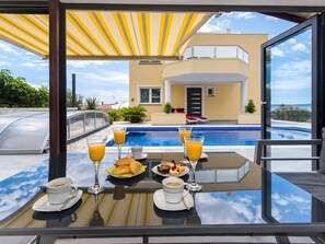 A dining table with breakfast items and drinks, overlooking a swimming pool and a modern building.